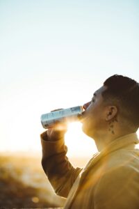 Person drinking water from a reusable bottle while resting in the shade on a hot day.